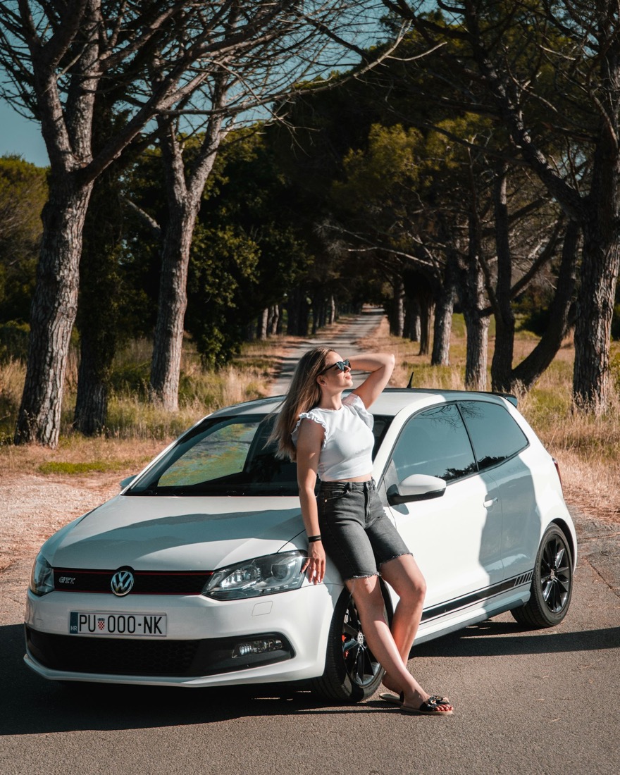 Jeune femme appuyée contre une voiture blanche, sur une route bordée de pins